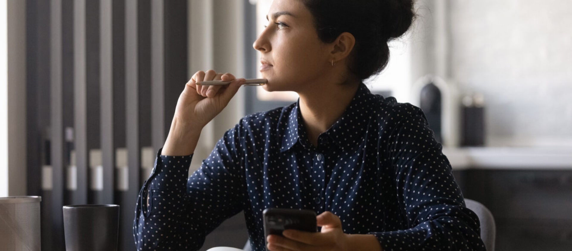 Woman looking out of the window thinking of a solution to a problem with blue shirt phone and pen and notebook